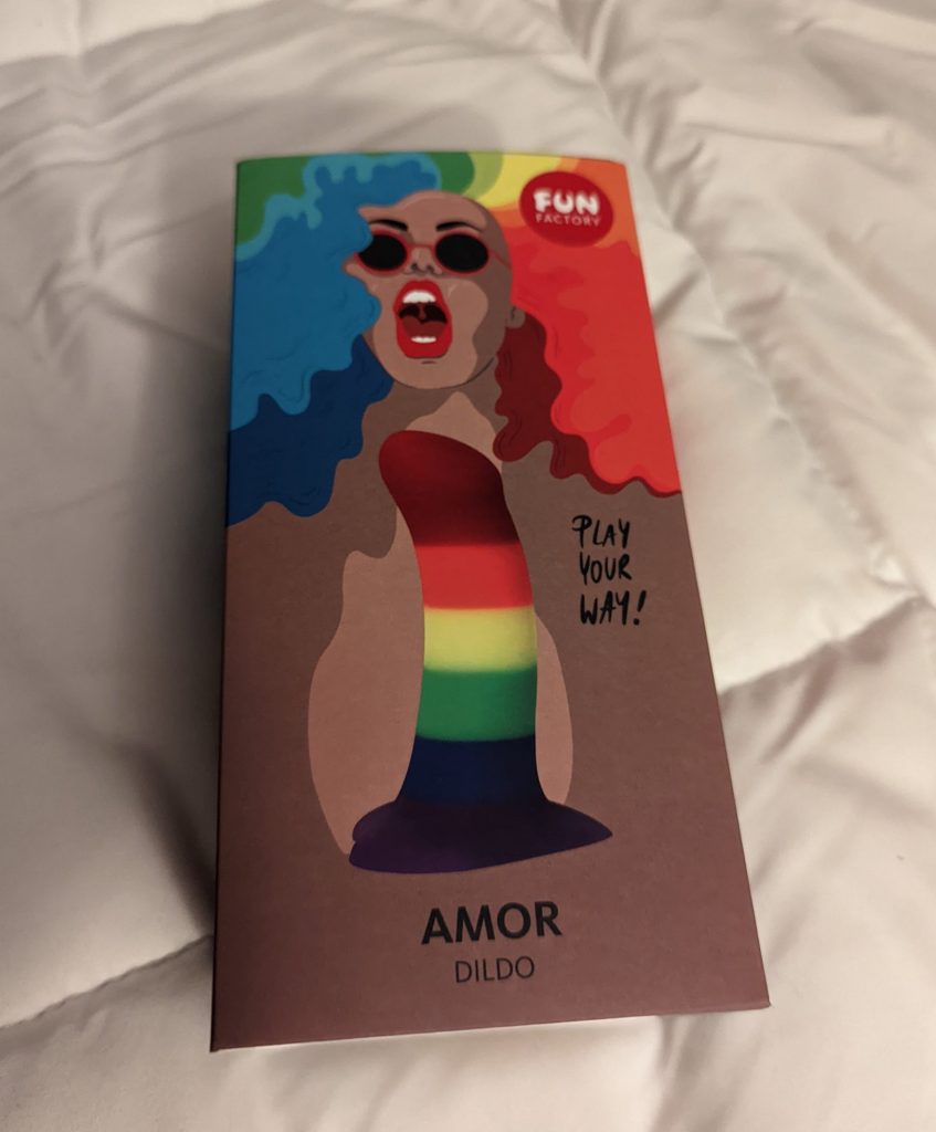 Rainbow amor dildo box with an image of the dildo and an illustration of a person with multicolored hair, sunglasses, and an open joyful mouth